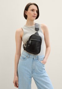 Sac en cuir noir en bandoulière avec une forme incurvée, doté d'une poche zippée à l'avant et d'une sangle réglable à motifs.