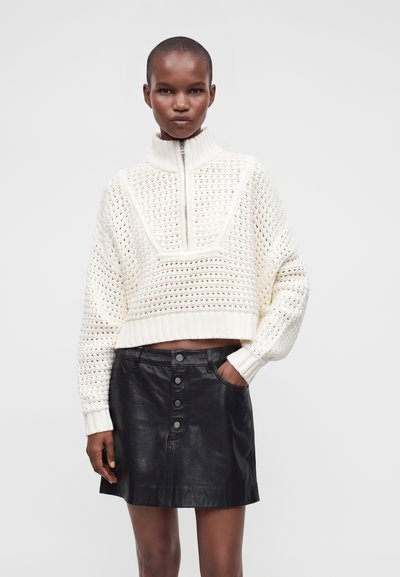 Femme portant un pull en maille texturé blanc avec une fermeture éclair mi-longue et une jupe en cuir noire à boutons sur le devant, debout avec une main dans la poche.