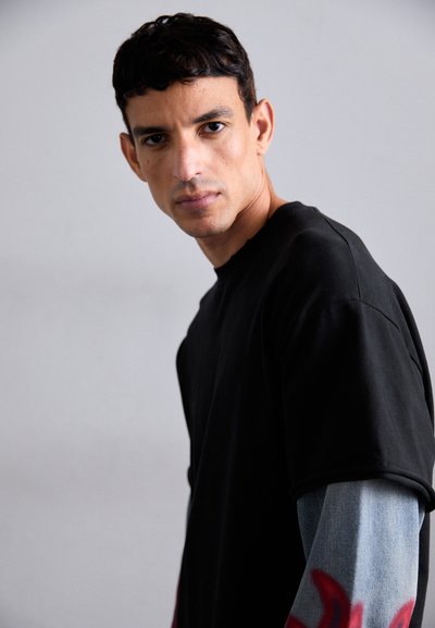 Tricou negru oversized cu mâneci scurte, purtat peste o cămașă de blugi bleu deschis cu mâneci lungi, care prezintă elemente decorative roșii.