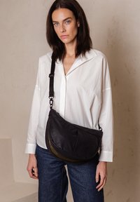 Borsa a tracolla nera con design curvo, tracolla regolabile e tessuto testurizzato. Abbinata a una camicia bianca con bottoni e jeans blu.