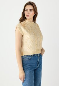 Gebreide, korte trui in lichtgoud met een hoge ronde halslijn, korte mouwen en een textuur van kabelpatronen. Gepaard met blauwe denim jeans.