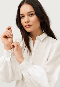 Witte linnen blouse met een knoopsluiting, voorzien van pofmouwen en elastische manchetten. Model is haar mouwen aan het verstellen.