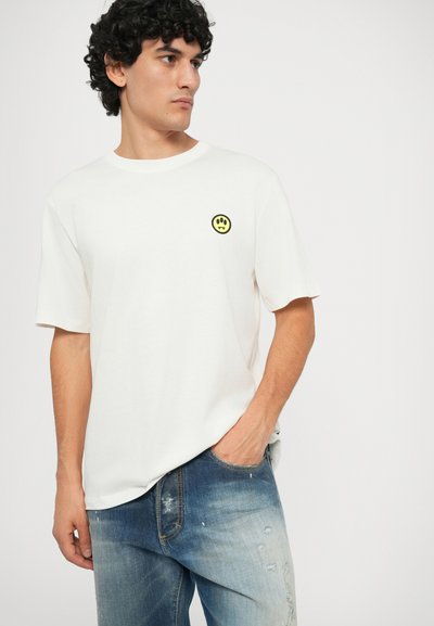Camiseta de algodón blanca con mangas cortas y escote redondo. Presenta un gráfico circular en negro y amarillo en el lado izquierdo del pecho. Combinada con jeans azules desgastados.