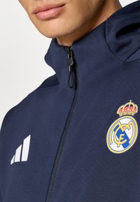 Navy Zip-Jacke mit hohem Kragen, einem kleinen Reißverschluss und weißen Adidas-Streifen. Zeigt das Real Madrid-Logo mit einer Krone.