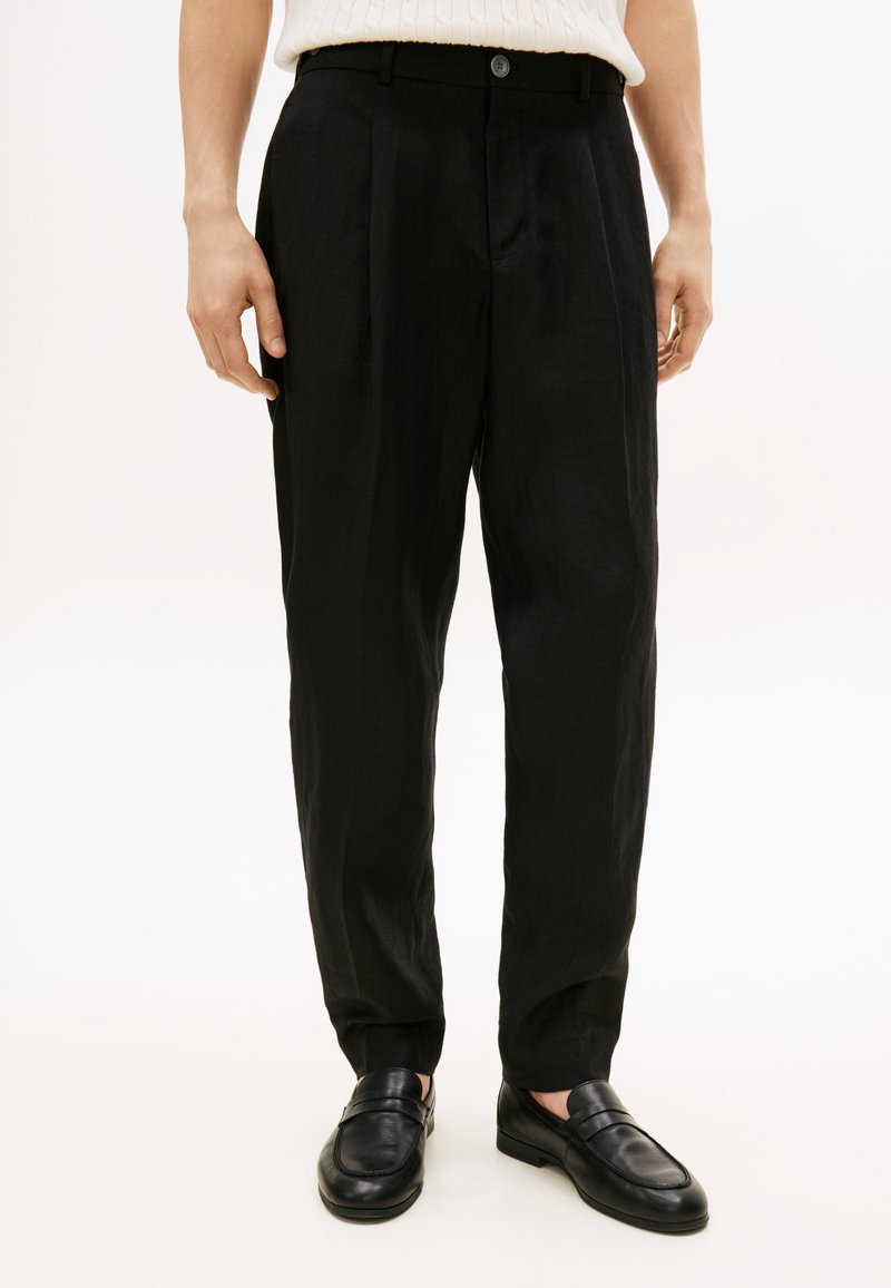 Άνδρας που φοράει μαύρο πλισέ παντελόνι και μαύρα δερμάτινα μοκασίνια, στέκεται μπροστά σε απλό φωτεινό φόντο.