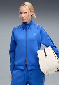 Chaqueta deportiva azul con cuello alto, cierre de cremallera y acentos blancos. Combinada con un bolso crema que tiene una cremallera y ribetes negros.