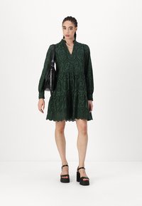 Green lace dress with a V-neck, long puff sleeves, tiered skirt, and scalloped hem. Accessorized with a black handbag and platform sandals.