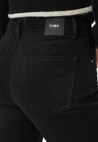 Jeans in denim nero con una superficie liscia, design minimalista e un'etichetta del marchio ben visibile sulla cintura; presenta accenti di cucitura discreti.