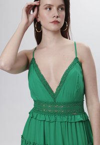 Vestido verde de tejido ligero que presenta un escote en V profundo, ribete de encaje, dobladillo con volantes y tirantes de espagueti ajustables.