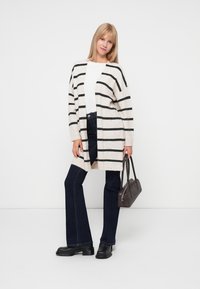 Beige Strickjacke mit schwarzen Streifen, offene Vorderseite, lange Ärmel, getragen über einem weißen Oberteil und dunklen Jeans, kombiniert mit einer kleinen braunen Handtasche und Stiefeln.