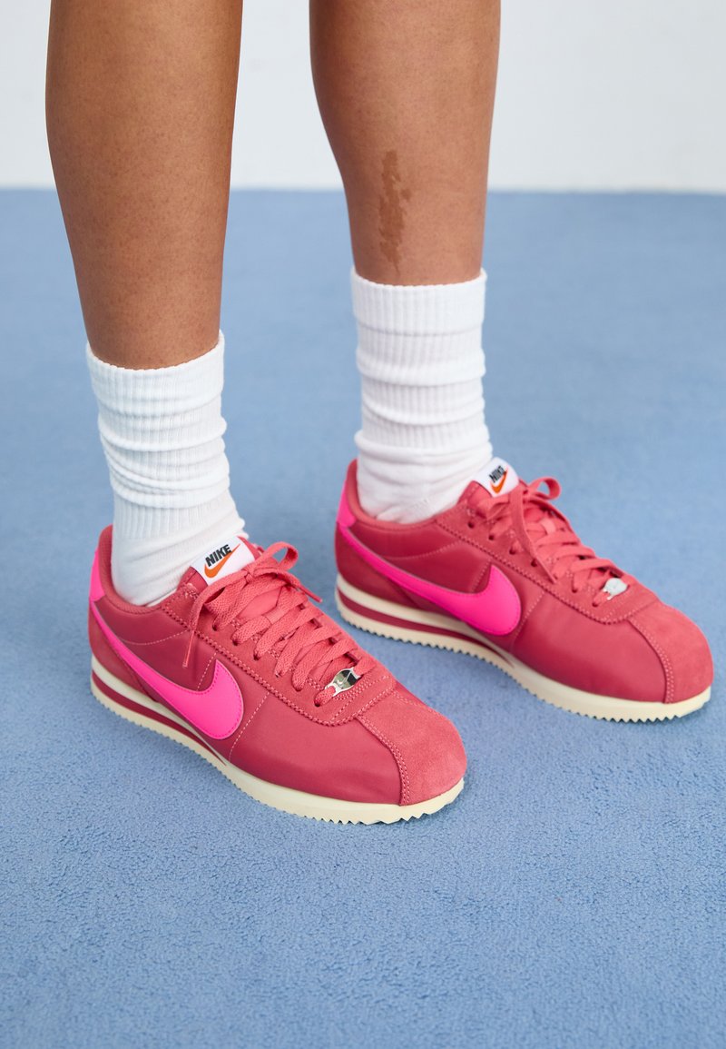 Rózsaszín Nike sportcipők, amelyek sima és texturált anyagok kombinációjával készültek, merész rózsaszín pipa és fehér fűző található rajtuk, fehér zoknikkal párosítva.