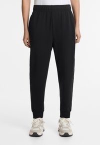 Black sweatpants with elastic waistband, tapered leg design, and side pockets. Paired with light gray sneakers featuring cream accents.