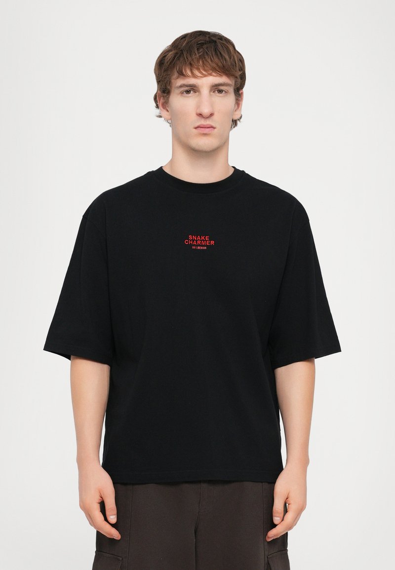 T-shirt en coton noir à manches courtes, arborant un petit texte brodé rouge "SNAKE CHARMER" sur la poitrine, avec un col rond.
