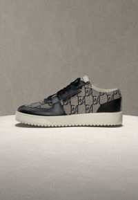 Baskets basses avec détails en cuir noir et tissu beige à motif de lettres foncées, semelle blanche, présentées sur une surface claire.