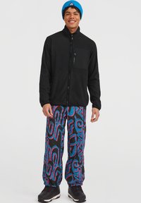 Giacca in pile nera con zip e tasca sul petto, abbinata a pantaloni neri con vivaci motivi astratti blu e rosa. Scarpe nere e berretto blu.