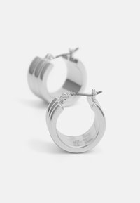 Hopeilla korvakoruilla, joissa on sileä, heijastava pinta ja kolme vaakasuoraa uurteista. Niissä on turvallinen salpa, joka helpottaa pukemista.