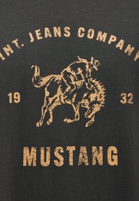 Grafika jezdce na bokujícím koni s oprýskaným textem "INT. JEANS COMPANY," "1932," a "MUSTANG" na tmavém látkovém pozadí.