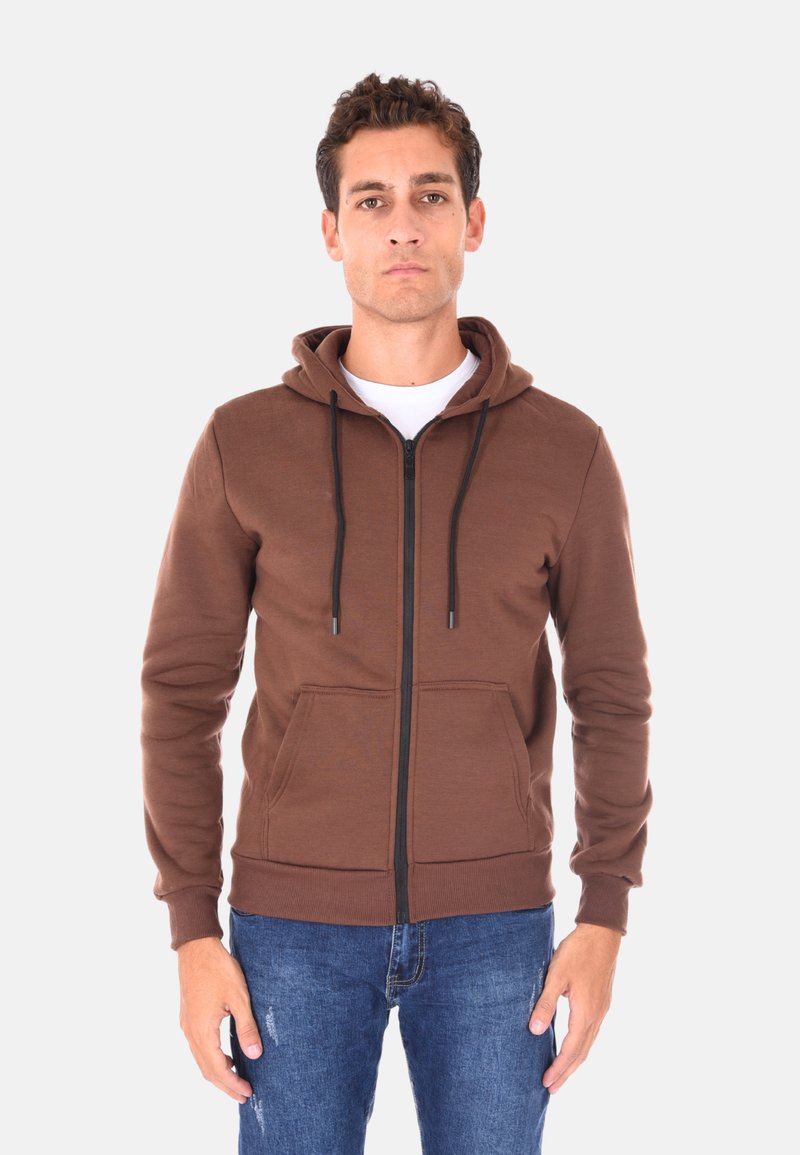 Felpa con zip marrone dotata di cappuccio con lacci, tasche frontali e polsini a coste. Indossata sopra una maglietta bianca e abbinata a jeans blu.