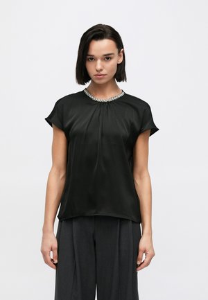 Svart satinblus med plisserad front och ärmlösa axelvaddar, prydd med en pärlbeklädd halsringning. Kombinerad med grå vida byxor.