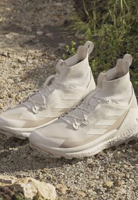 Beżowe buty sportowe z wysokim, dzianinowym kołnierzem, teksturowaną cholewką i gumową podeszwą. Posiadają minimalne sznurowanie i logo na pięcie.