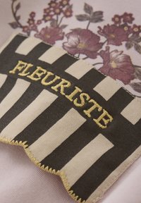 Tygmärke med blommönster i nyanser av lila, med en svartvit rand och broderad text "FLEURISTE" i guldtråd.