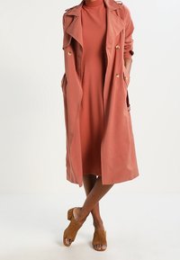 Lång, lätt korallfärgad trenchcoat med guldknappar, lager-på-lager över en matchande klänning. Bärs med bruna öppna muletofflor.