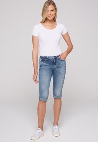 Pantalones cortos bermuda de mezclilla azul claro con puños enrollados, que presentan acentos de texto bordados en la parte frontal, combinados con una camiseta ajustada blanca.