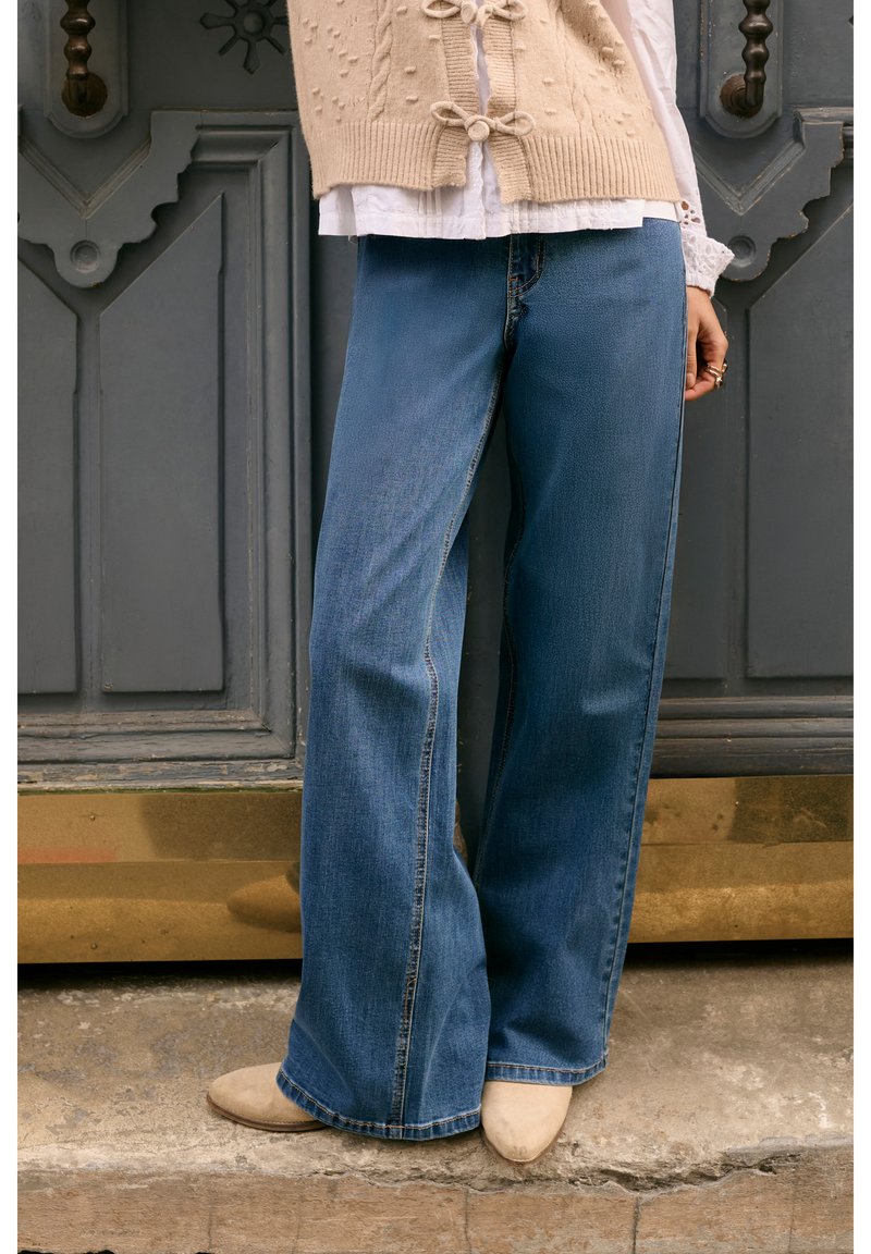 Denimflared jeans i mellemblå, med høj talje, rå kant og let falmning. Sammen med en beige strikket vest og cremefarvede sko.