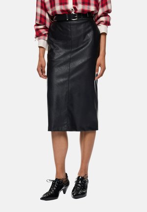 Donna che indossa una gonna midi in pelle nera con cintura, camicia rossa a quadri e scarpe nere con tacco e lacci, in piedi davanti a uno sfondo semplice.