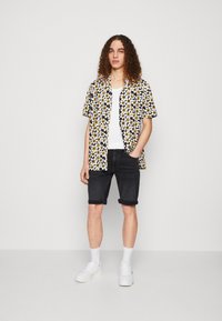Camisa negra de manga corta con un patrón floral en blanco y amarillo, combinada con pantalones cortos de denim negros enrollados y zapatillas blancas.