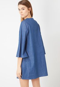 Joven mujer con cabello castaño claro que lleva un vestido de mezclilla azul suelto con mangas acampanadas de tres cuartos, mirando por encima de su hombro.