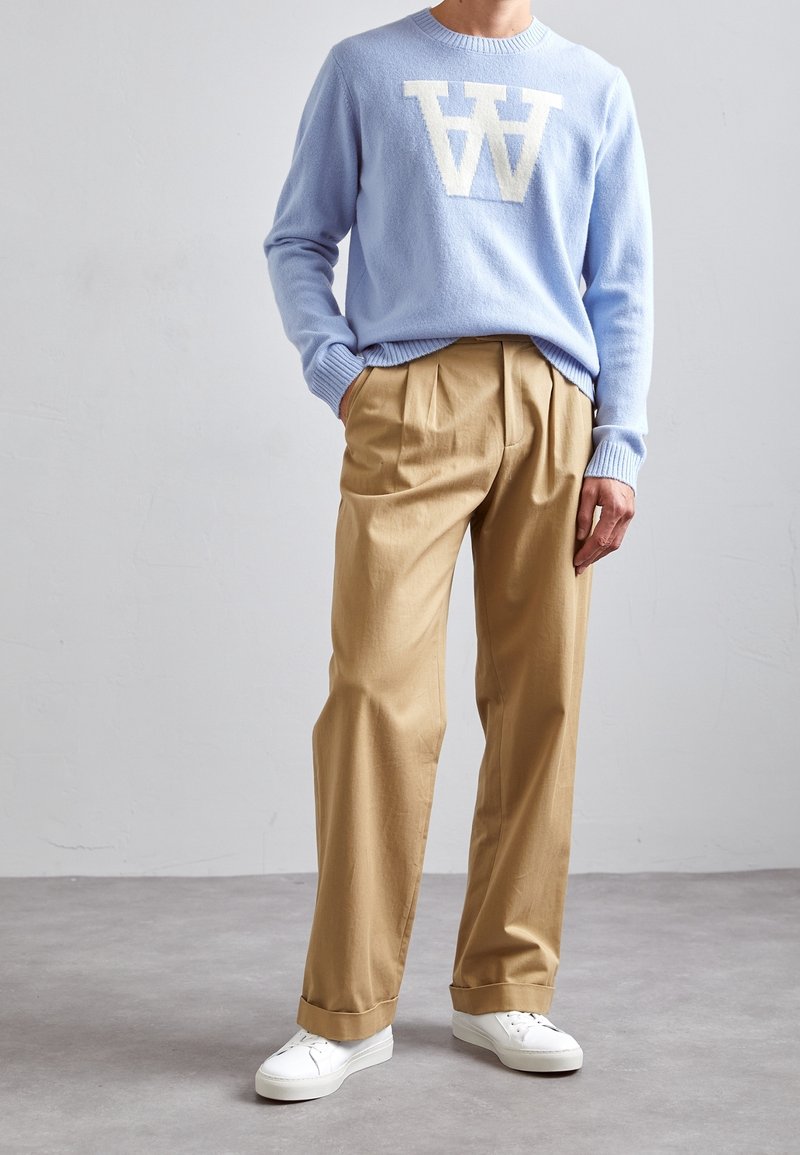 Pull bleu clair avec un motif en lettre blanche, associé à un pantalon beige plissé. Des baskets blanches complètent la tenue.