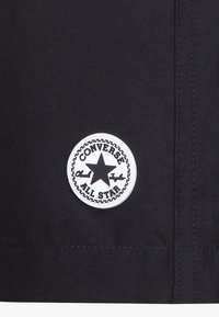 Tissu noir présentant un patch rond blanc avec une étoile noire et le texte "CONVERSE ALL STAR" autour, posé sur une couture piquée.