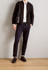 Homme portant une veste noire, une chemise blanche, un pantalon bleu marine foncé, des mocassins en cuir noirs, et portant un sac bandoulière noir, debout sur un sol en bois.