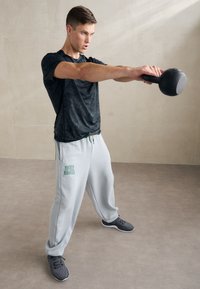 Šedé tepláky s logem "Under Armour", černé strukturované tričko a šedé sportovní boty. Osoba zvedající kettlebell v neutrálním prostředí.