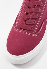 Scarpa da ginnastica in tela color bordeaux con suola in gomma bianca. Presenta lacci piatti e cuciture visibili lungo i lati. Il puntale testurizzato è ben evidente.