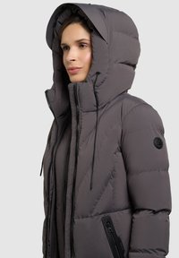 Harmaan sävyinen toppatakki, jossa on irrotettava huppu, diagonaalinen tikkaus, musta vetoketju ja vetoketjulliset sivutaskut.