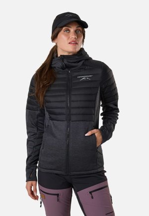 Femme portant une casquette noire, une veste matelassée noire et gris foncé, et un pantalon d'extérieur violet avec des poches zippées, posant avec une main dans la poche de la veste.