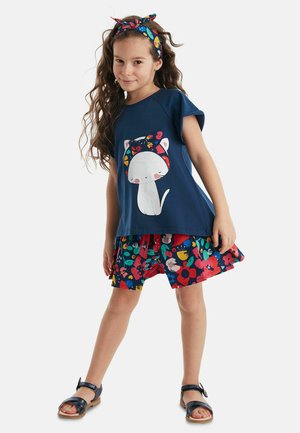 Camiseta de color azul marino con un gráfico de gato, combinada con pantalones cortos de estampado floral colorido. Incluye una diadema a juego. Las sandalias son azules con hebilla.