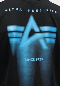Μαύρο ύφασμα με μεγάλο μπλε στυλιζαρισμένο λογότυπο «A», οριζόντιες ρίγες, κείμενο «ALPHA INDUSTRIES» πάνω από το λογότυπο και «SINCE 1959» κάτω από το λογότυπο.