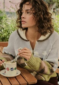 Knit sweater with a zip front; pastel colors including light blue, green, and brown; ribbed cuffs; floral-patterned ceramic mug on a wooden table.