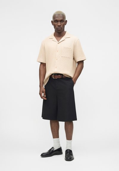 Άνδρας με κοντά ξανθά μαλλιά, ντυμένος με μπεζ κοντομάνικο πουκάμισο, μαύρα φαρδιά σορτς, άσπρες κάλτσες και μαύρα loafers, στάσιμος με το ένα χέρι στην τσέπη.