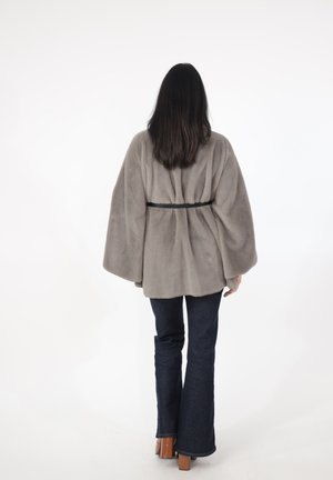 Kvinde med langt mørkt hår iført en grå pelsjakke med bælte, mørke vide jeans og brune sko med blokhæl, stående med ryggen til.