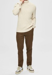 Beige kabelgebreide coltrui gecombineerd met olijfgroene broeken en crèmekleurige sneakers met crèmekleurige en bruine accenten.