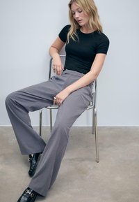 Žena sedící na kovové židli, oblečená v černém přiléhavém tričku, šedých širokých kalhotách, černých mokasínech a s několika prsteny na prstech.