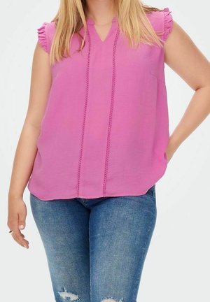 En kvinde iført en ærmeløs pink bluse med flæser på skuldrene og blå revnede jeans, der står med den ene hånd på hoften og den anden afslappet.