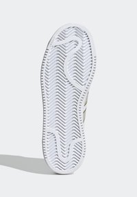 Bílá podrážka tenisky s vlnitým vzorem, zdobená embosovaným logem a hladkým, texturovaným okrajem.