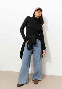 Un col roulé noir, un pantalon en denim bleu surdimensionné à jambes larges, une chemise rayée nouée à la taille, des chaussures en imprimé léopard brun, une casquette rose.