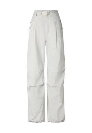 Vrouw met blond haar en zonnebril, draagt een zwart geknoopt cropped shirt, losse witte broek en witte sneakers, staand tegen een witte achtergrond.