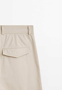 Section d'une jupe en tissu beige montrant une poche arrière avec rabat, des passants de ceinture et des détails de couture sur un fond blanc.